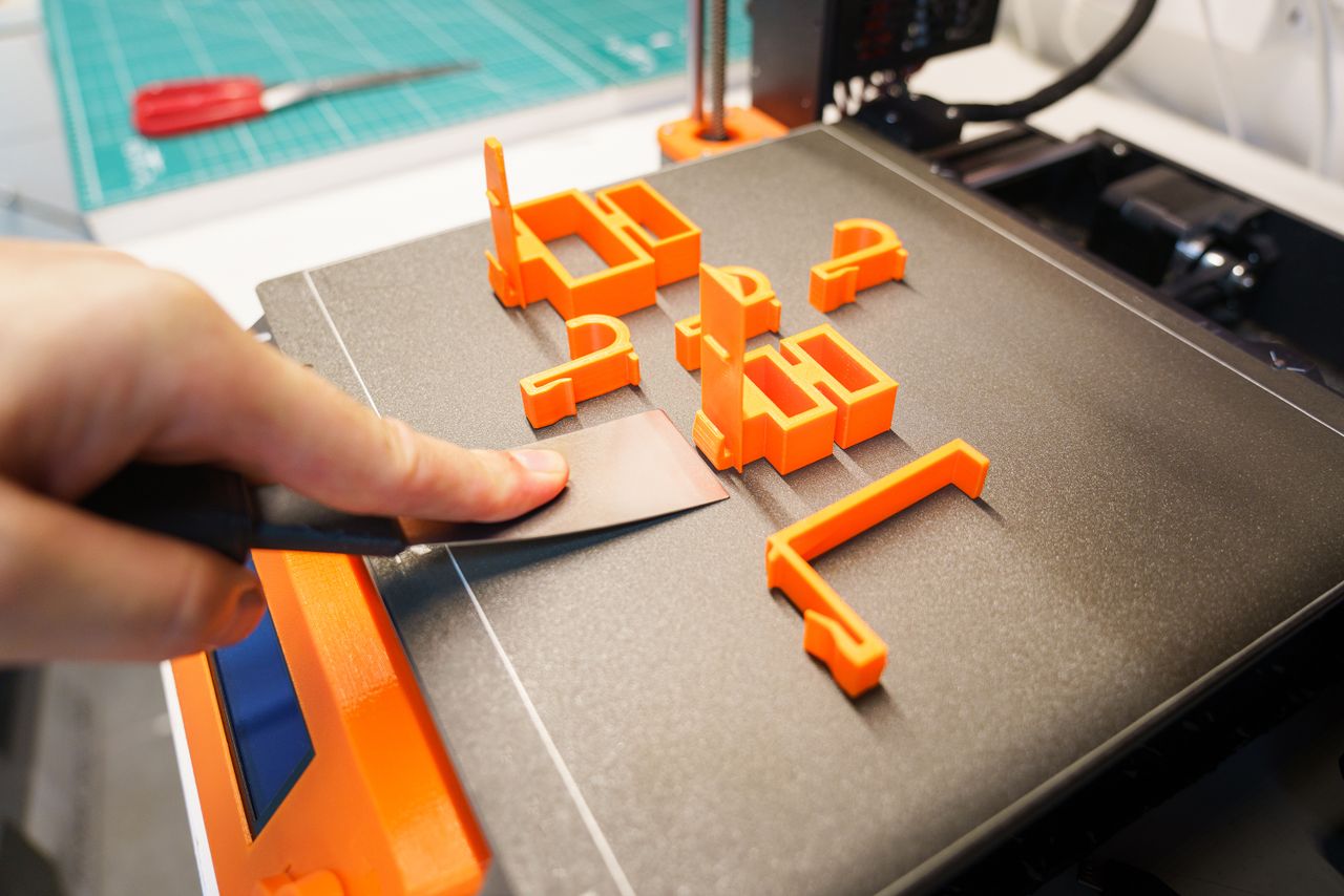 services-03 A hand carefully removes an orange 3D printed object from a printer bed using a spatula indoors.