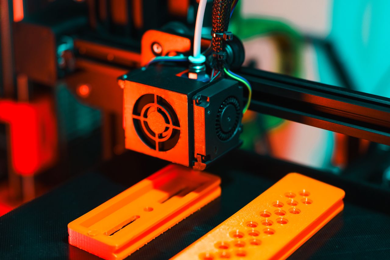 services-02 Detailed view of a 3D printer in action, extruding orange plastic components.