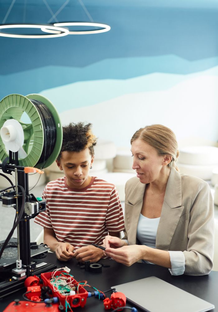 Concentrated female in casual outfit sitting with ethnic boy at table near laptop and working on project together in light room